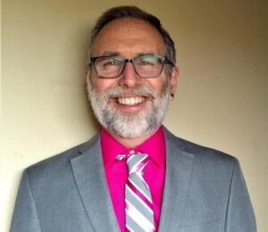 Rev. Phillip Schulman wearing a gray suit, red shirt and tie.